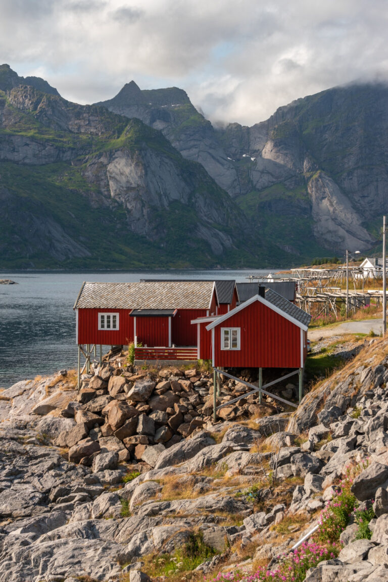 Red wooden cabins on a rocky shore by the water, with mountains behind